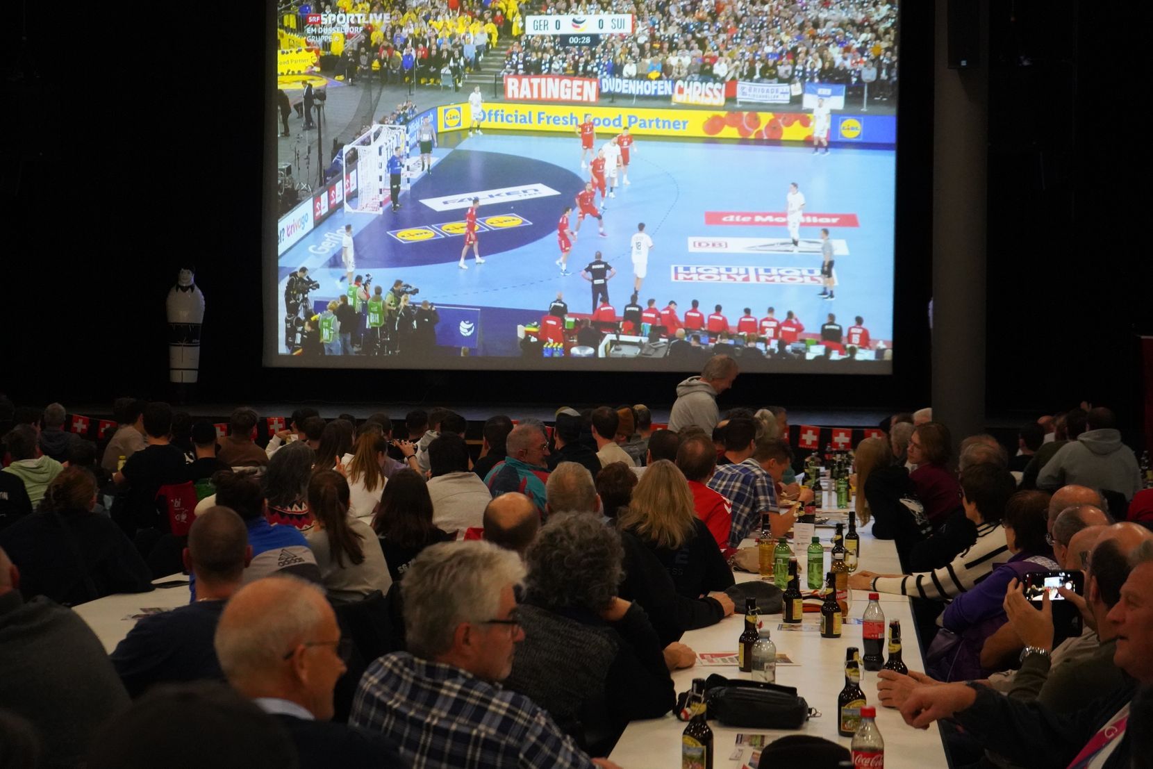 Das Publikum schaut gebannt auf den Bildschirm, wo die ersten Sekunden des Handball-EM-Eröffnungsspiels zwischen der Schweiz und Deutschland laufen.